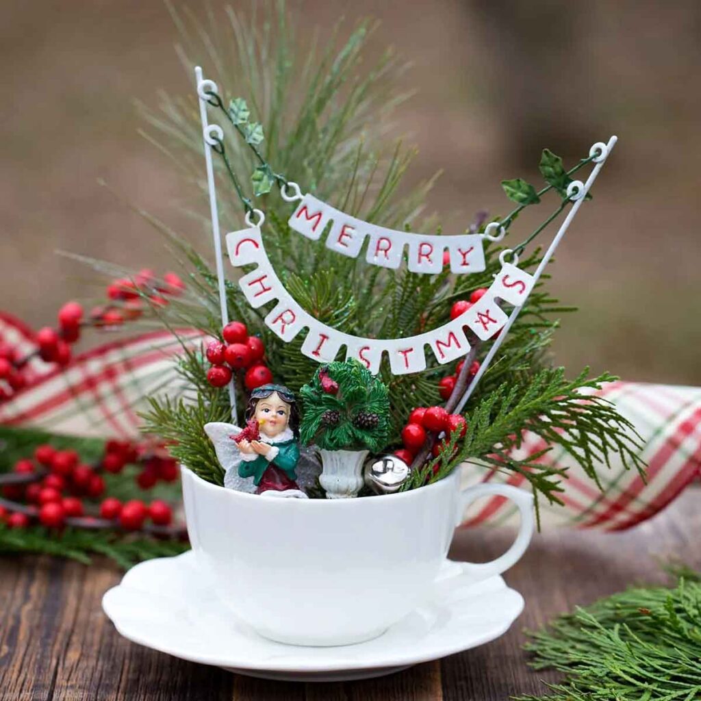 A white teacup displays a charming Christmas craft for teens, filled with greenery, red berries, a small angel figurine, and a mini banner reading "Merry Christmas." The cozy setup rests on a wooden surface with a plaid cloth backdrop.