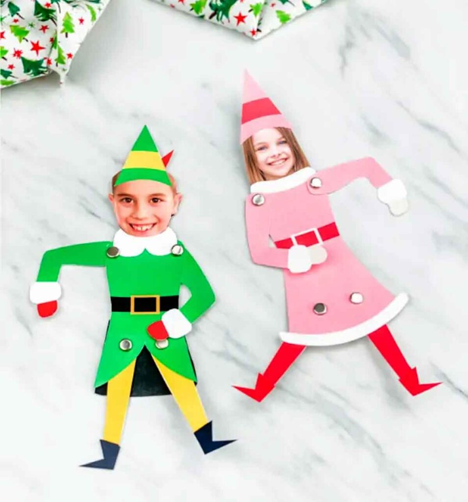 Two paper elf crafts on a marble surface, featuring cut-out photos of children's faces. The left elf wears green and yellow with a pointy hat, and the right elf wears pink and red with a similar hat. Wrapped gifts are partially visible.