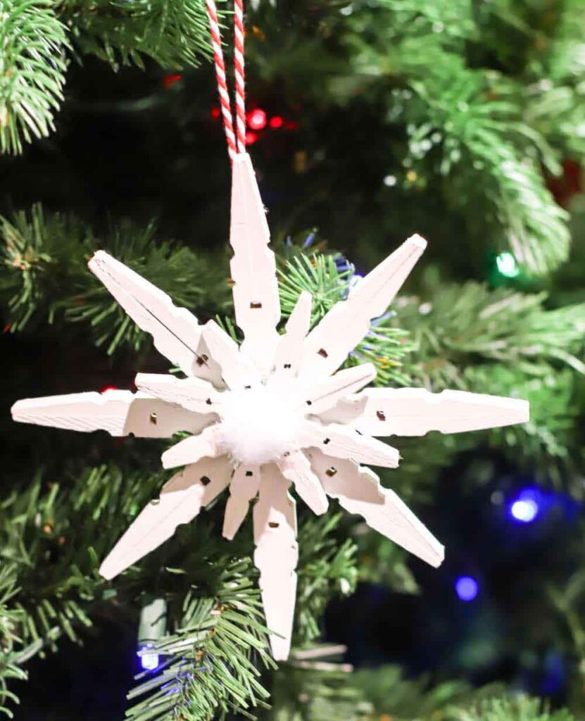 A star-shaped ornament made from white clothespins hangs on a Christmas tree adorned with colorful lights. Ideal for Christmas crafts for teens, the ornament is held by a red and white striped string.