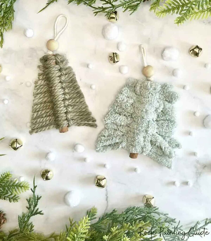 Two handcrafted Christmas tree ornaments made of green yarn and wooden beads are laid on a white surface. Ideal for christmas crafts for teens, the scene is decorated with green foliage, small white pom-poms, and gold jingle bells, creating a festive atmosphere.