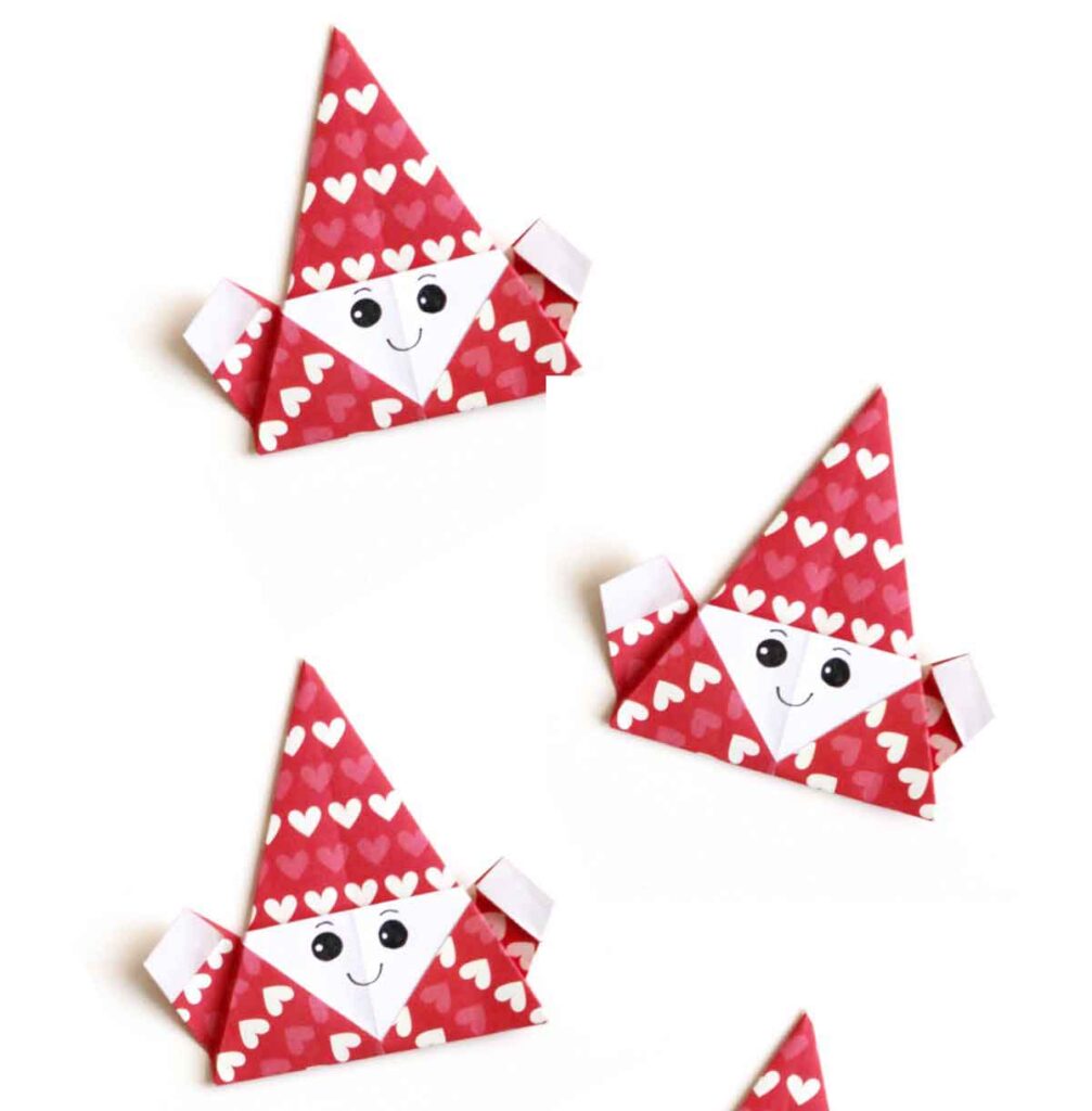 Four origami elf faces crafted with red and white paper, perfect for Christmas crafts for teens. Each charming face features a triangular red hat, cheerful eyes, a smile, and heart accents on a simple white backdrop.