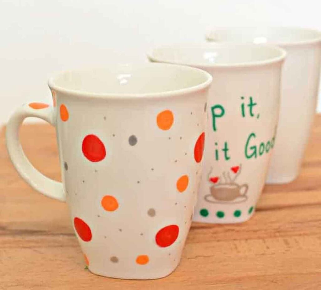 Three ceramic mugs are arranged in a diagonal line on a wooden surface, perfect for Christmas crafts for teens. The front mug boasts orange and gray polka dots, the middle features text with a teacup illustration, and the back mug stands plain white.