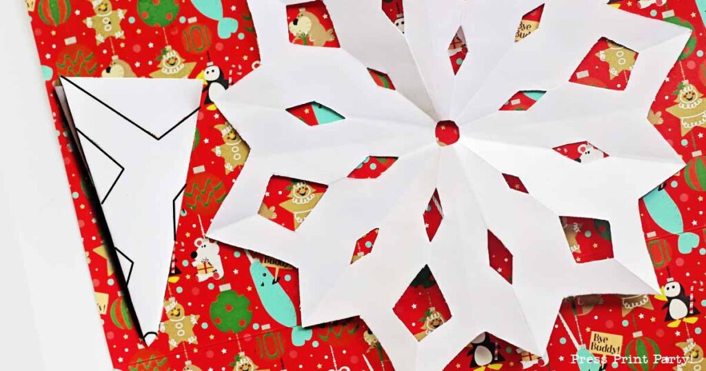 A large white paper snowflake, crafted from a template, is placed on festive red gift wrap adorned with snowmen and bear illustrations. A folded triangular piece of paper rests to the side, hinting at its role in creating the snowflake masterpiece.