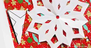 A large white paper snowflake, crafted from a template, is placed on festive red gift wrap adorned with snowmen and bear illustrations. A folded triangular piece of paper rests to the side, hinting at its role in creating the snowflake masterpiece.