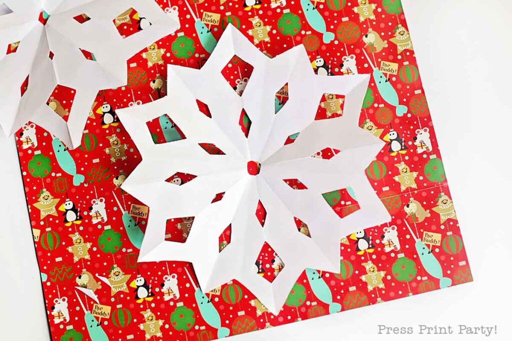 A white paper snowflake with a red center is placed on red Christmas-themed wrapping paper decorated with cartoon penguins, snowmen, gift boxes, and ornaments.