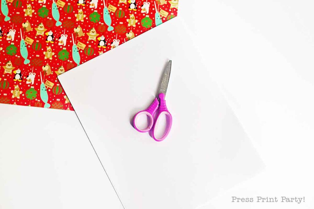 Purple scissors on a white sheet of paper, lying next to a colorful piece of Christmas-themed wrapping paper with patterns of trees, ornaments, and stars on a white surface.