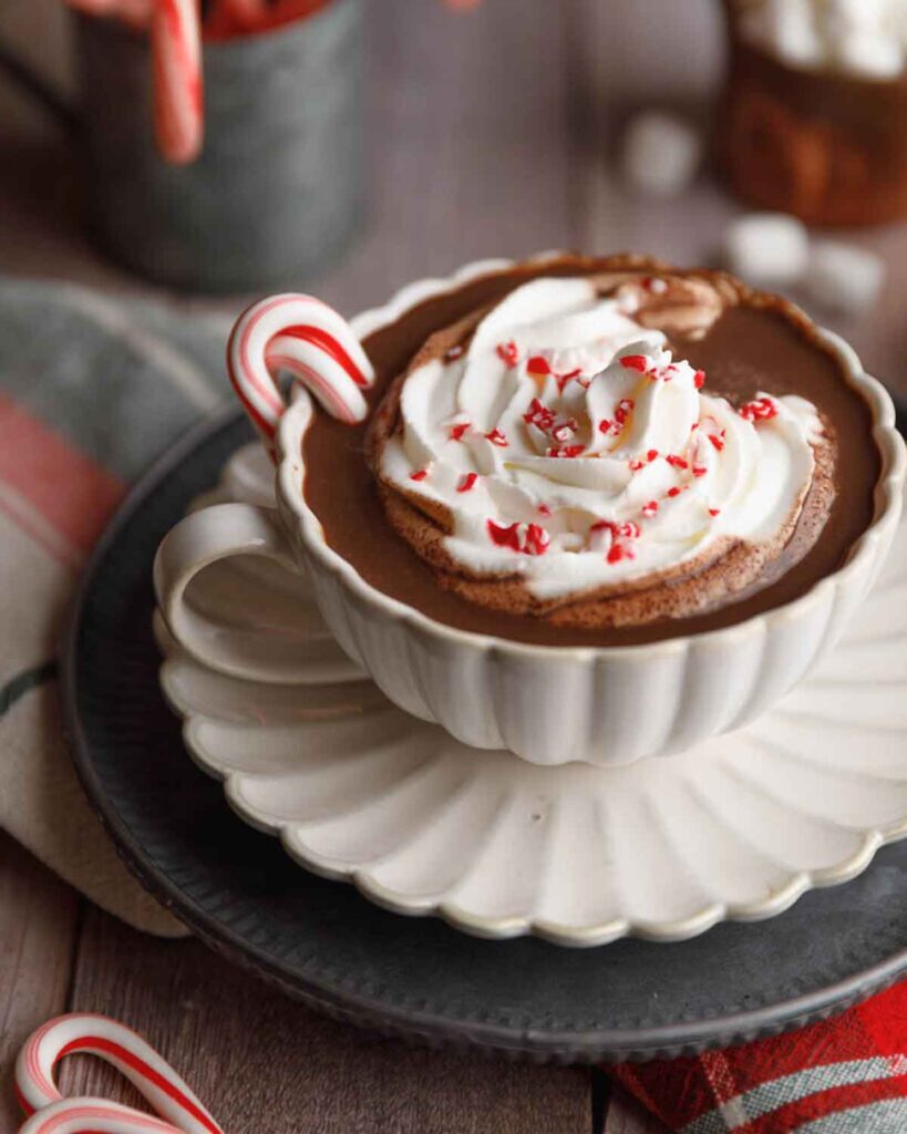 A cup of hot chocolate topped with whipped cream and red sprinkles sits on a scalloped saucer. A candy cane is hooked on the cup's handle. In the background, blurred candy canes and marshmallows are visible.