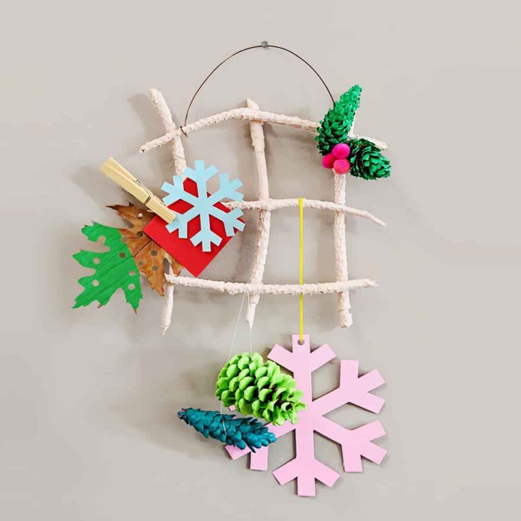 A grid made of twigs displays various colorful Christmas crafts for teens, including paper snowflakes, leaves, and pine cones. The crafts are attached with a clothespin and string, creating a playful and festive decoration against a neutral background.