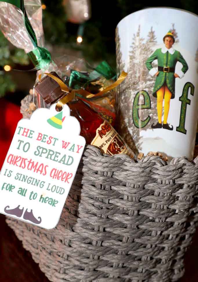 A festive basket containing a cup with an elf design, a small bottle of alcohol, and a holiday tag. The tag reads, "The best way to spread Christmas cheer is singing loud for all to hear.