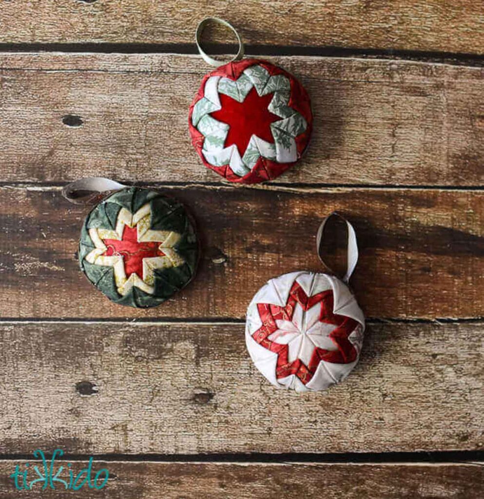 Three quilted Christmas ornaments with star patterns hang on a rustic wooden background, perfect as Christmas crafts for teens. Designed in red, green, and white, each ornament features intricate fabric folds and ribbon loops for hanging.