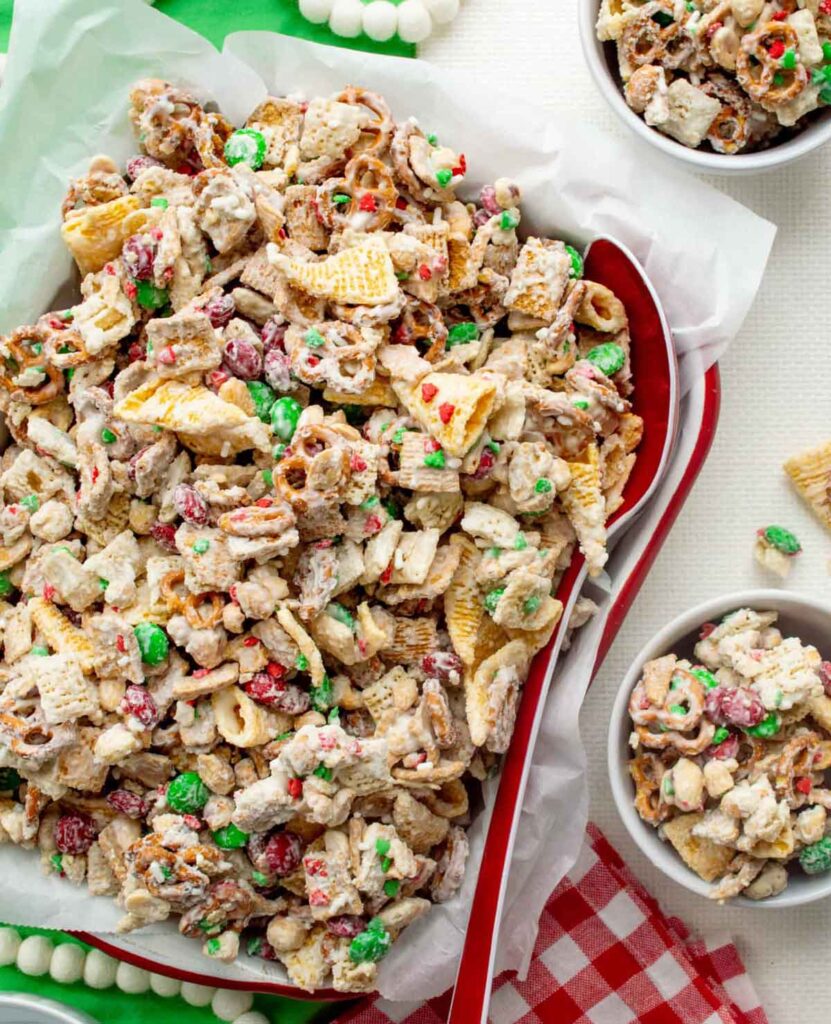 A tray filled with a festive snack mix, including cereal pieces, pretzels, peanuts, and red and green candies, coated in a white, sugary glaze. Two small bowls with the same snack mix are next to the tray on a red-checkered cloth.