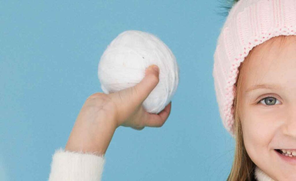 A child wearing a pink knitted hat holds a white snowball in one hand. The background is a bright blue, and the child is smiling, wearing a cozy white sweater. Only half of the child's face is visible in the image.