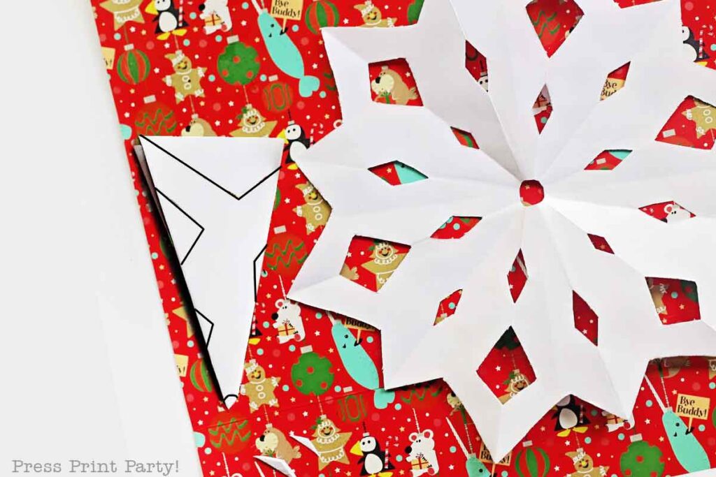 A white paper snowflake, crafted from templates, and an origami piece rest atop red Christmas-themed wrapping paper adorned with festive designs, including penguins and ornaments.
