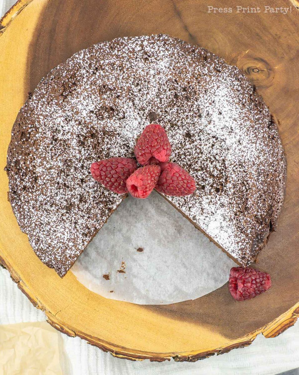 A round, 4-ingredient flourless chocolate cake is dusted with powdered sugar and topped with fresh raspberries. A slice has been removed, unveiling its moist interior. The cake elegantly rests on a wooden serving board.