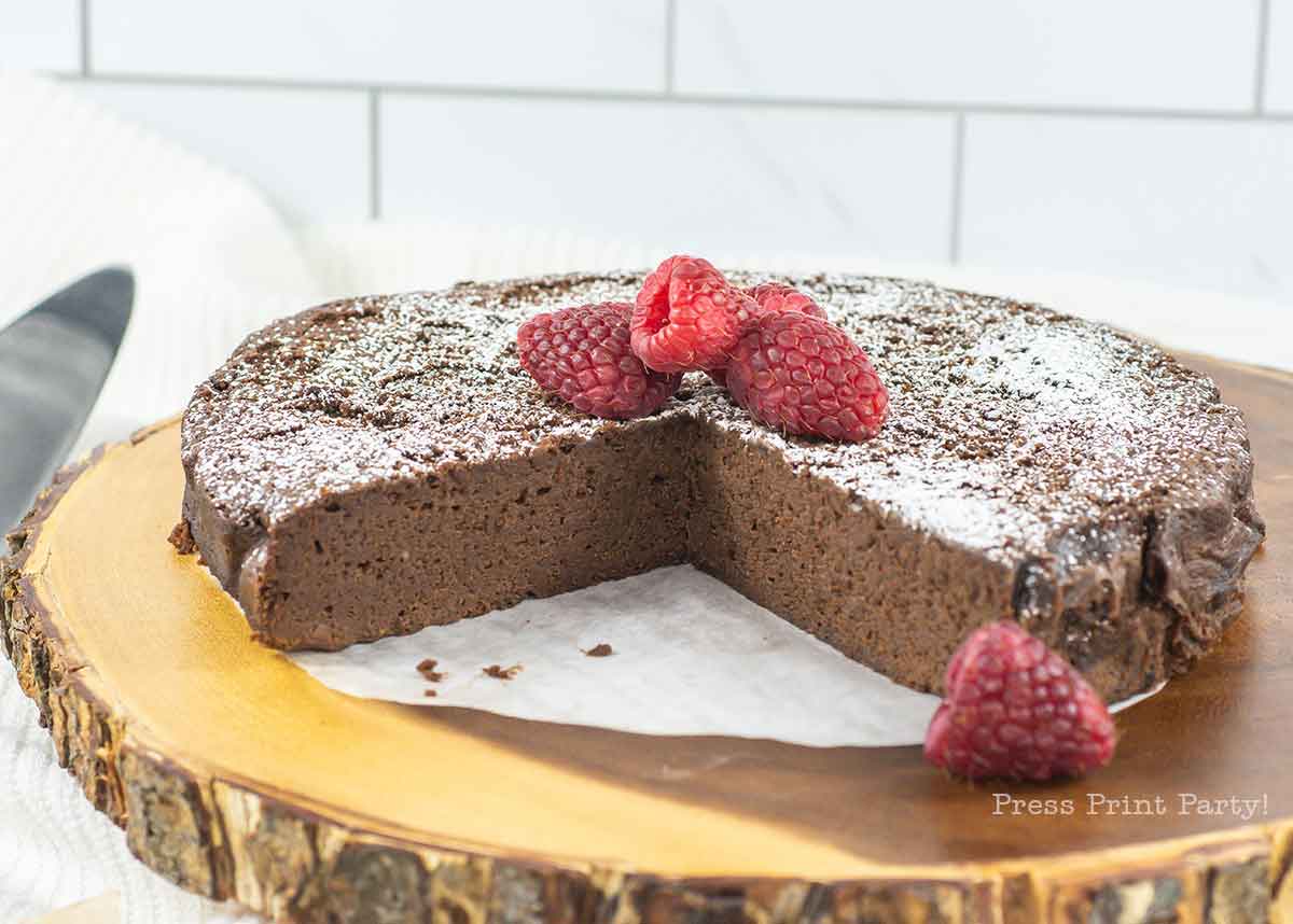 A sliced 4-ingredient flourless chocolate cake dusted with powdered sugar sits on a wooden platter. It's topped with fresh raspberries, with one slice visibly removed, showcasing its dense texture. The background features a light, tiled wall.