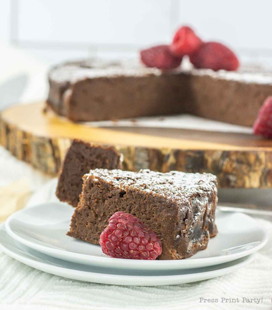 A slice of 4 ingredient flourless chocolate cake dusted with powdered sugar sits on a plate, garnished with fresh raspberries. In the background, a whole cake rests on a wooden board, missing just one tempting slice.