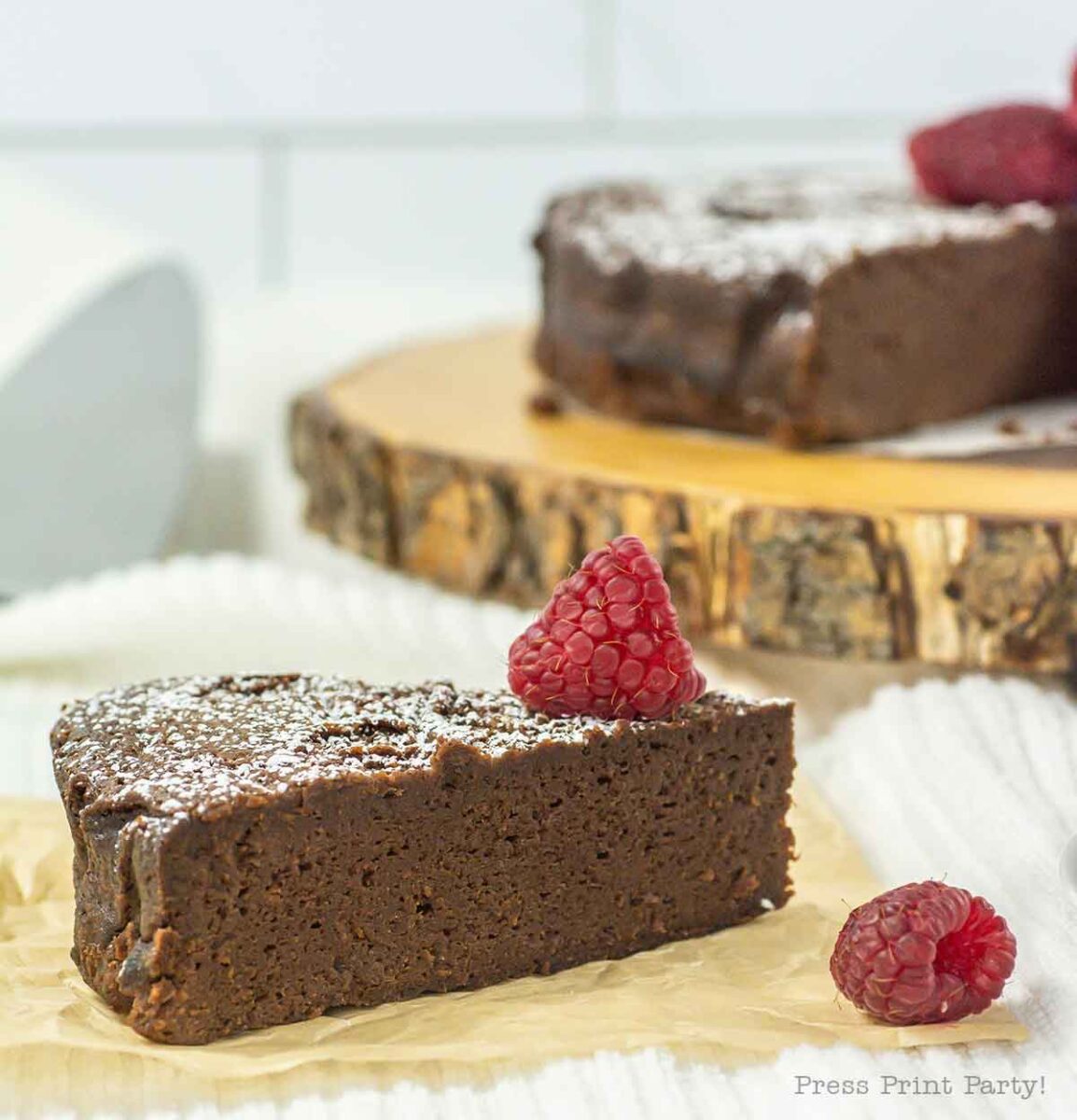 A slice of rich, 4-ingredient flourless chocolate cake is garnished with a fresh raspberry and a dusting of powdered sugar. The slice sits on parchment paper, while the rest of the indulgent cake rests invitingly on a wooden board in the background.