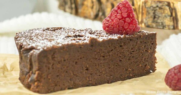 A slice of 4-ingredient flourless chocolate cake, topped with a raspberry and dusted with powdered sugar, sits elegantly on parchment paper. In the background, a tree bark decoration adds a touch of rustic charm.
