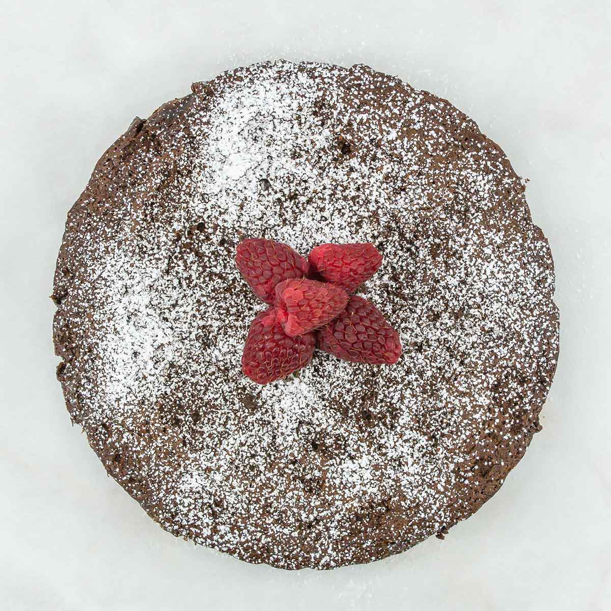 Overhead view of a 4-ingredient flourless chocolate cake dusted with powdered sugar, topped with a cluster of fresh raspberries in the center, set against a light background.