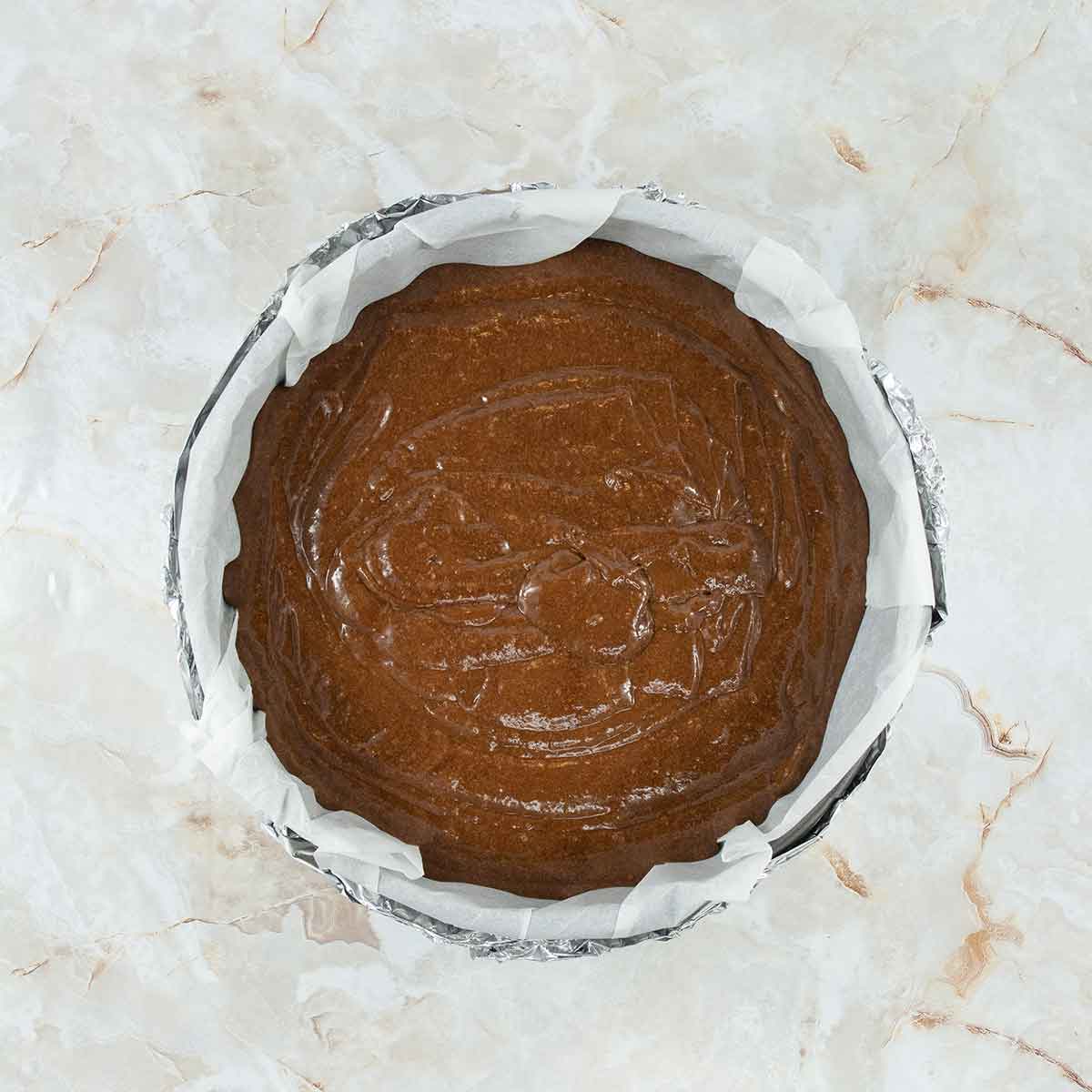 A round baking tin lined with parchment paper is filled with smooth batter for a 4 ingredient flourless chocolate cake, ready for baking. The background is a marble surface.
