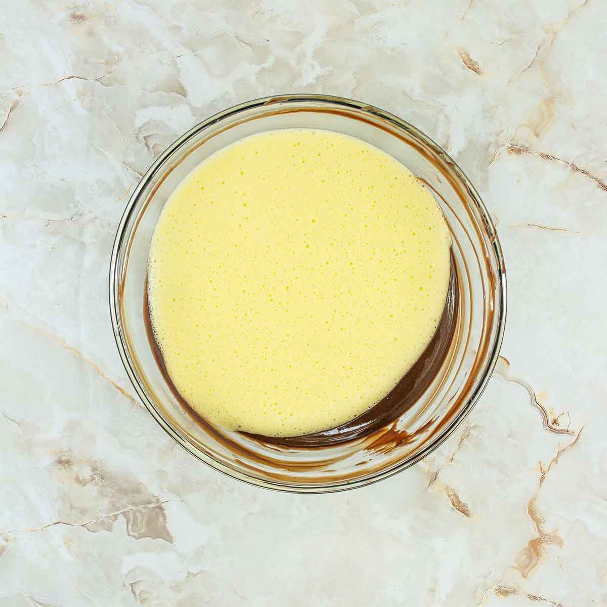 A glass bowl on a marbled surface holds the beginnings of a 4-ingredient flourless chocolate cake, with creamy yellow batter partially blended with darker chocolate swirls.