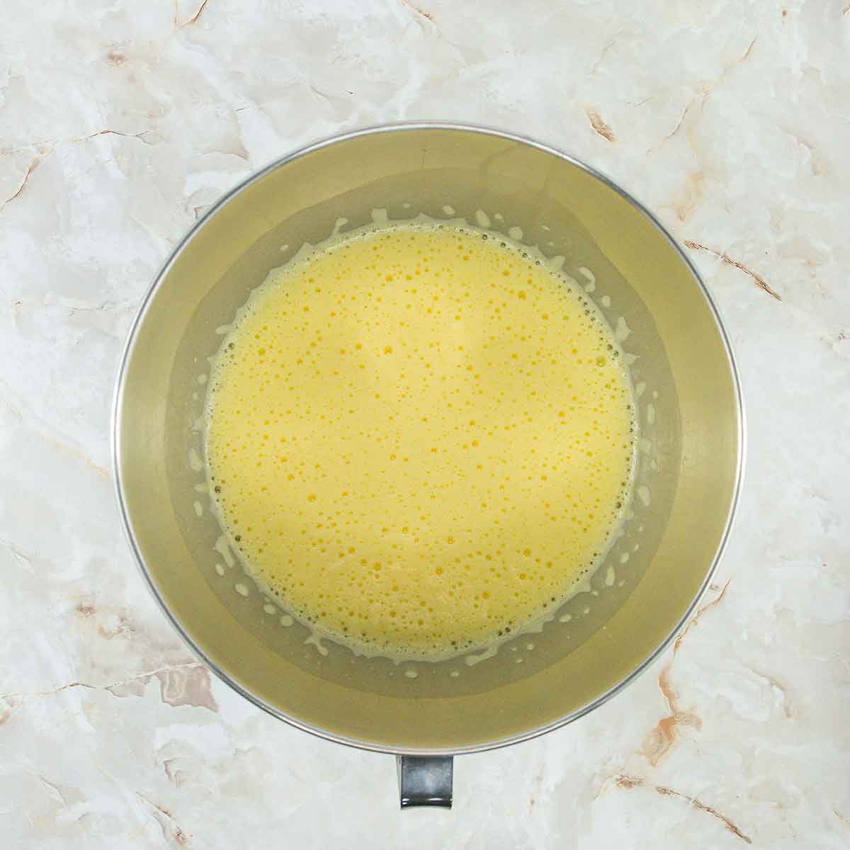 A stainless steel mixing bowl filled with frothy, light yellow liquid rests on a marble countertop, poised for perfection. The smooth blend hints at its role in crafting a 4-ingredient flourless chocolate cake.