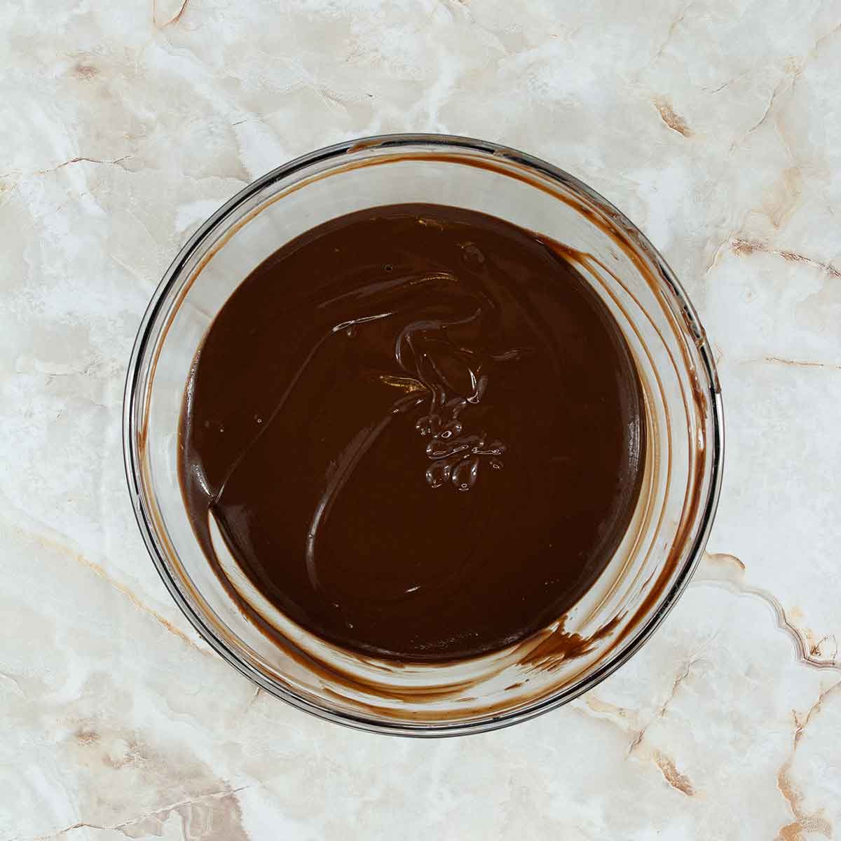 A glass bowl filled with smooth, melted chocolate, perfect for crafting a 4 ingredient flourless chocolate cake, sits on a marbled countertop. The glossy texture of the chocolate forms enticing streaks along the bowl's edges.