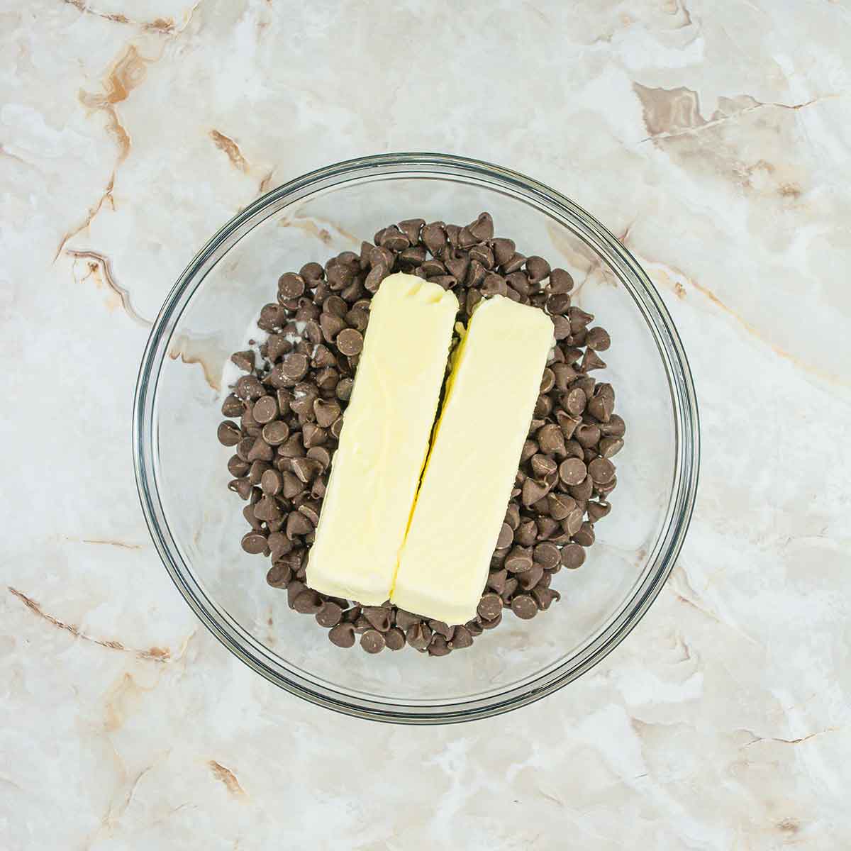 On a marbled countertop, a glass bowl brims with chocolate chips and two sticks of butter, ready to craft a delectable 4-ingredient flourless chocolate cake.