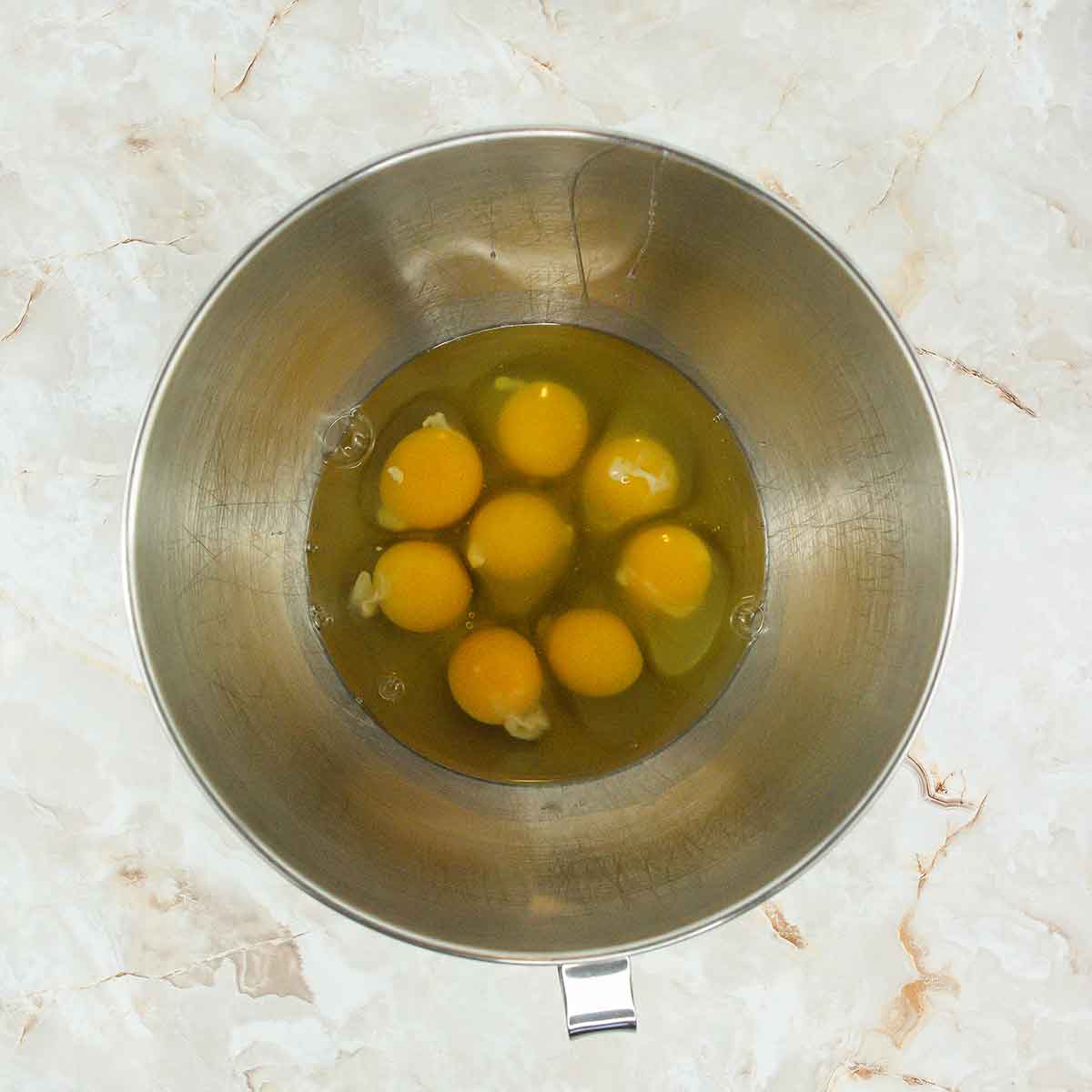 A stainless steel mixing bowl containing seven cracked eggs, yolks intact, sits on a light marble countertop—perfect for crafting a 4 ingredient flourless chocolate cake.