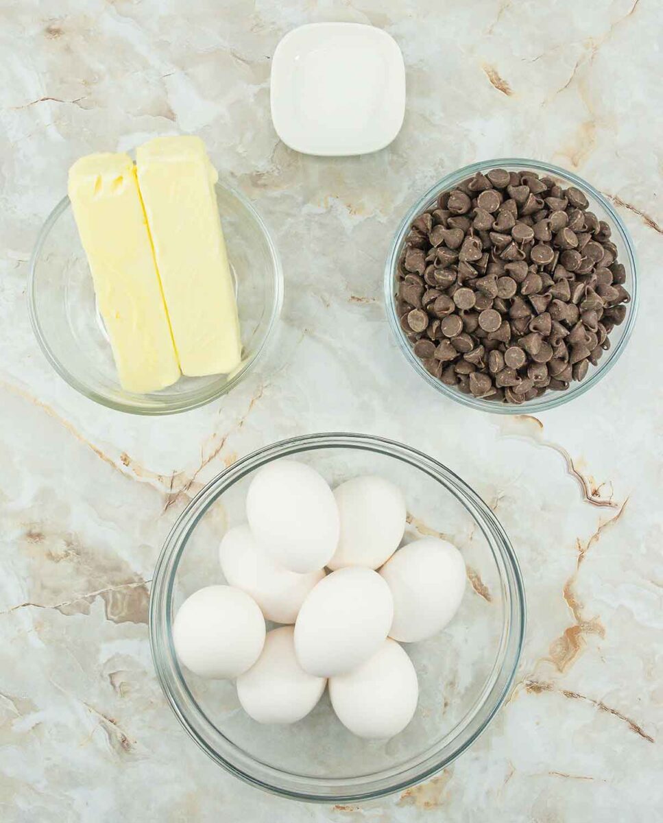 Four bowls on a marble surface contain essential ingredients for a delicious 4-ingredient flourless chocolate cake. The bowls hold butter, chocolate chips, eggs, and a small amount of salt, promising an indulgent treat with minimal effort.