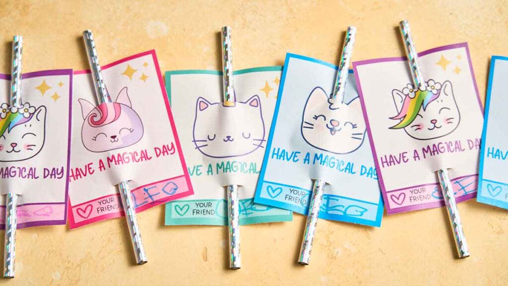 Colorful cards with cute cat drawings, each showcasing a unique design and the message "Have a Magical Day." Attached silver straws add charm to these whimsical pencil valentines, all beautifully displayed on a light yellow surface.