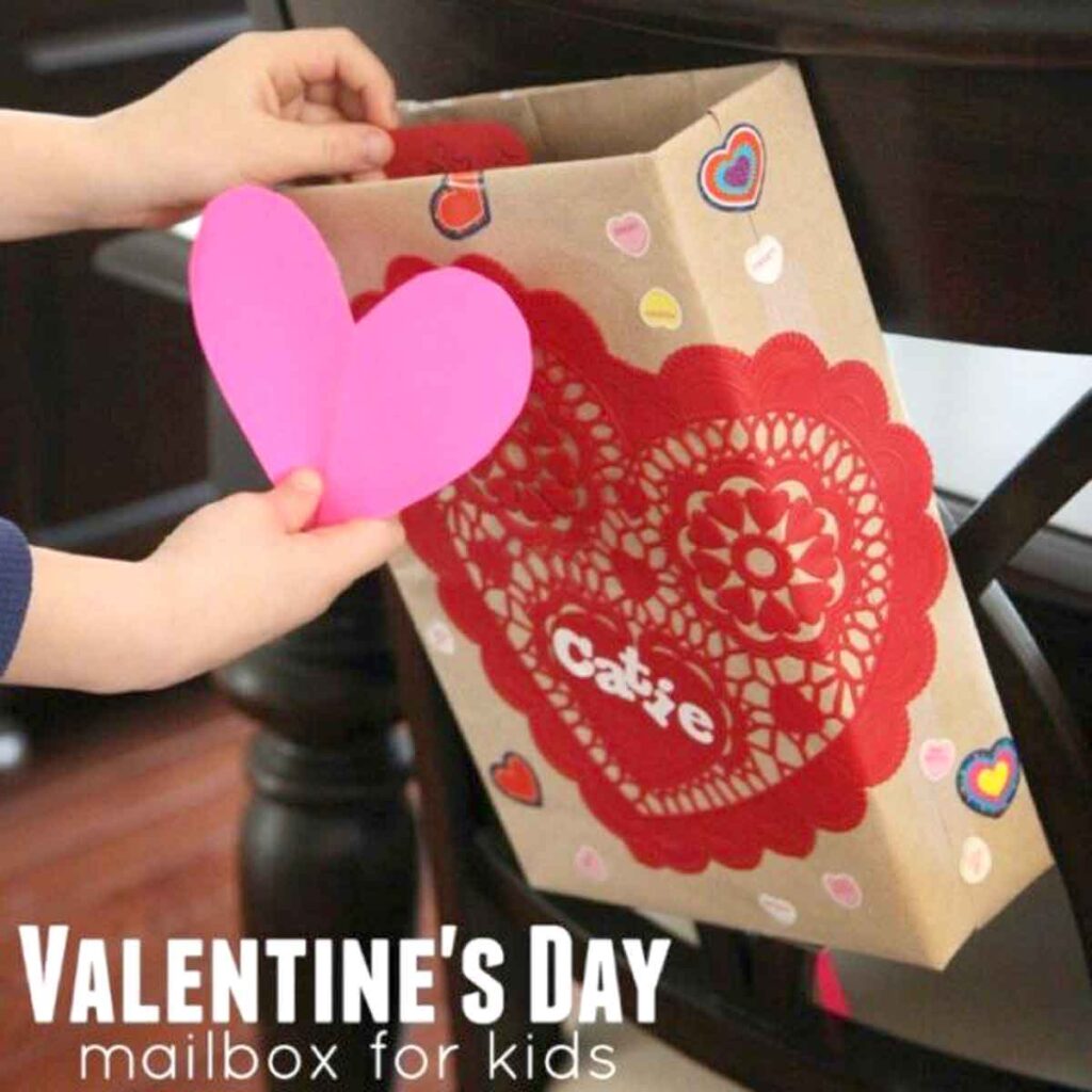 Child's hands placing a pink heart into a Valentine's Day mailbox, adorned with a red paper doily and heart stickers. This creative valentine box idea for school is attached to a chair, with "Valentine's Day mailbox for kids" inscribed at the bottom.