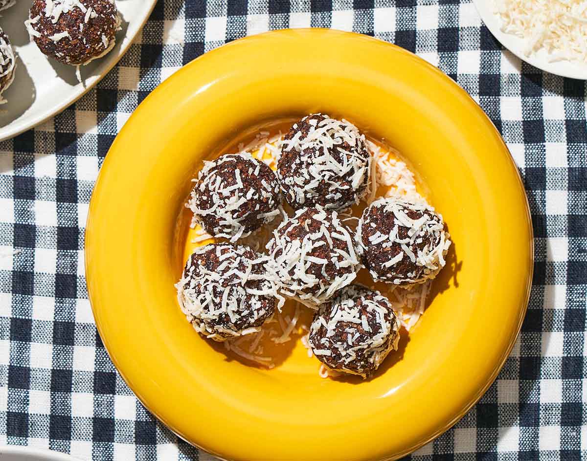 Seven chocolate balls covered in shredded coconut are arranged on a bright yellow plate, making delightful healthy Valentine snacks for the classroom. The plate is set on a black-and-white checkered tablecloth, adding a touch of whimsical charm.