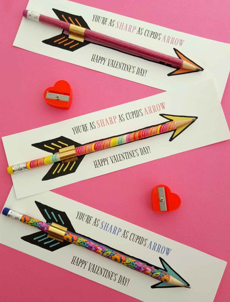 Three colorful pencil valentines resembling arrows are placed on cards with "You're as sharp as Cupid's arrow. Happy Valentine's Day!" written on them. The cards rest on a pink surface, surrounded by small, red heart-shaped sharpeners.