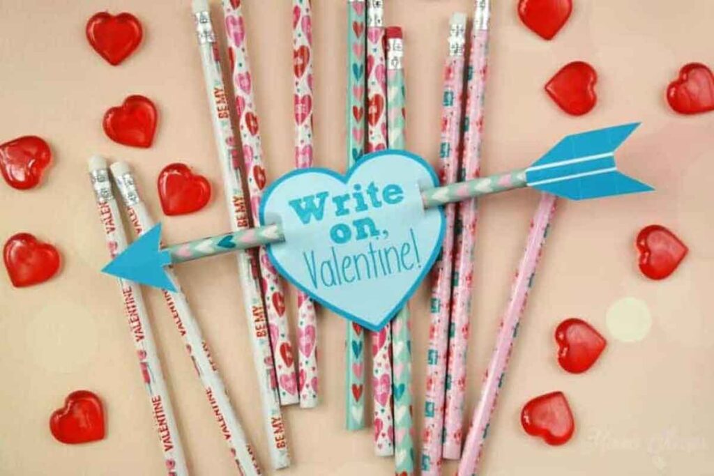 A collection of pencil valentines is showcased with a blue heart and arrow in the center reading "Write on, Valentine!" Red heart-shaped candies are scattered around on a light background, adding a sweet touch.
