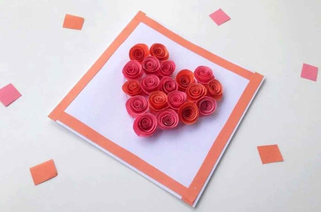 A handmade card featuring a heart shape made of rolled paper roses in red and orange hues. The card has an orange border and is surrounded by scattered orange paper squares on a white surface.