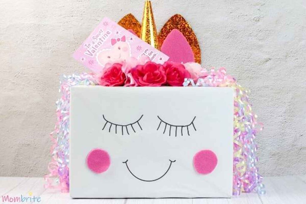 A decorated box resembling a unicorn, with pink cheeks, closed eyes, and a smiling mouth. This valentine box idea for school features glittery golden and pink ears, surrounded by colorful ribbons and flowers. A Valentine card is placed on top.