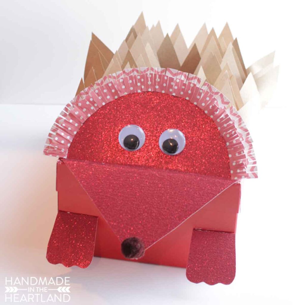 A red, glittery hedgehog craft perfect for valentine box ideas for school, made from paper with googly eyes, a black pom-pom nose, and brown paper spikes. The body boasts a textured red glitter finish and a paper ruffle. "Handmade in the Heartland" is penned in the corner.