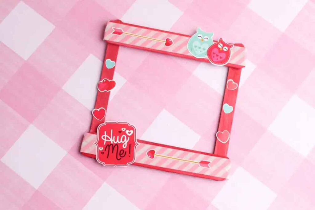 A decorative photo frame made of pink and red popsicle sticks, adorned with heart shapes and two colorful owls, perfect for Valentine paper crafts. A sign with the text "Hug Me!" is attached to one corner against a pink checkered background.
