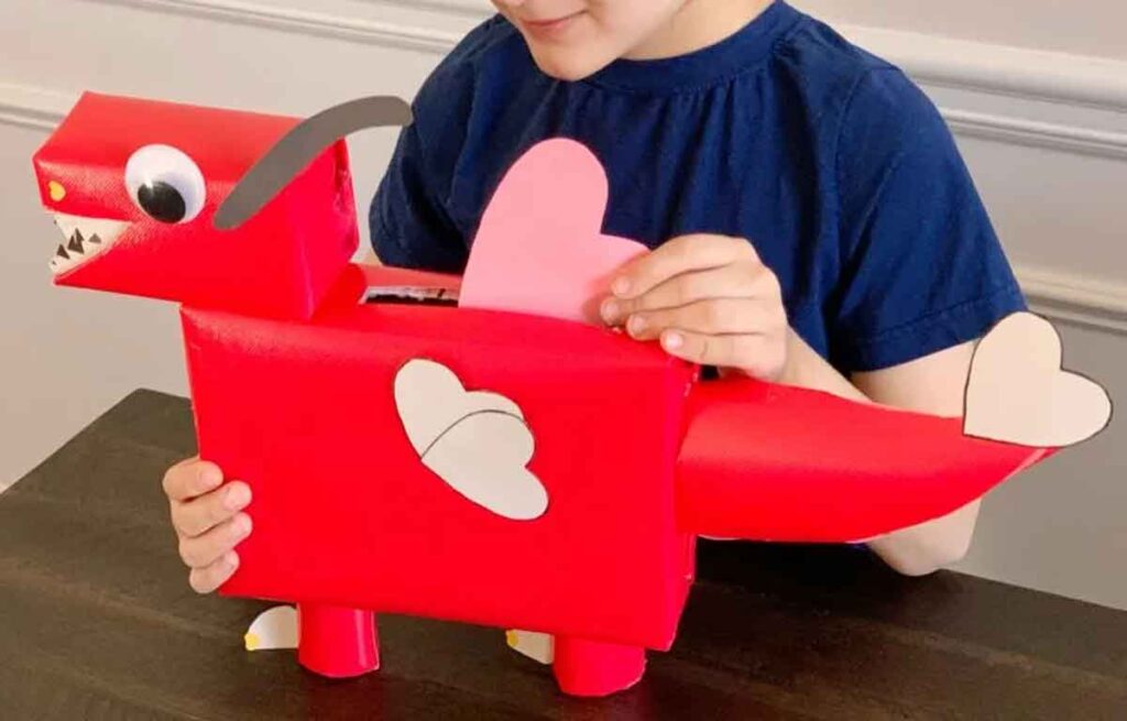 A child proudly holds a charming red dinosaur-shaped Valentine's box adorned with heart decorations—an imaginative valentine box idea for school. As the dinosaur stretches its long neck, the child eagerly inserts a pink heart into its back, all set against a dark surface.