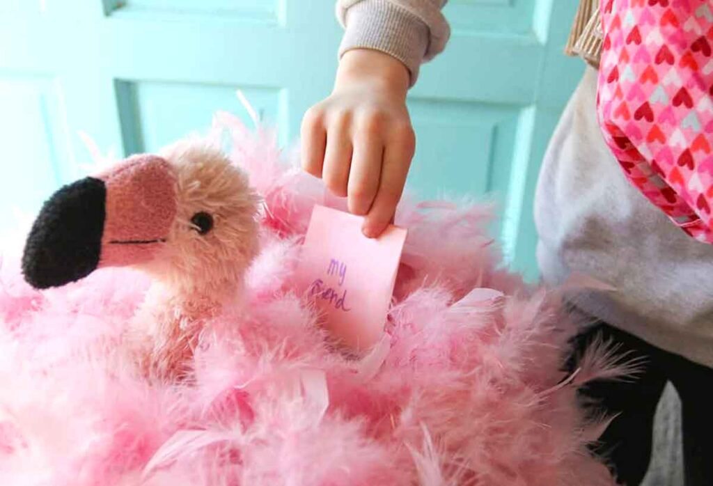 A child places a pink note labeled "my friend" into a fluffy, plush flamingo toy covered with pink feathers—a creative take on valentine box ideas for school. The background features a light teal door, and the child is wearing a heart-patterned accessory.