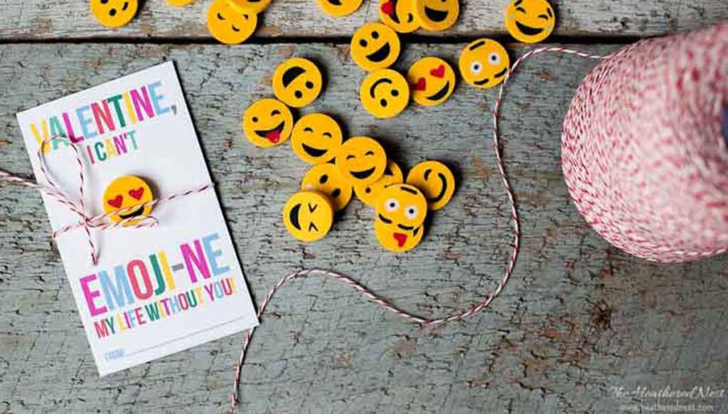 A collection of yellow emoji faces on a wooden surface next to a red and white twine spool and pencils. A card reads, "Valentine, I can't emoji-ne my life without you," with emojis printed on it, capturing the spirit of pencil valentines perfectly.