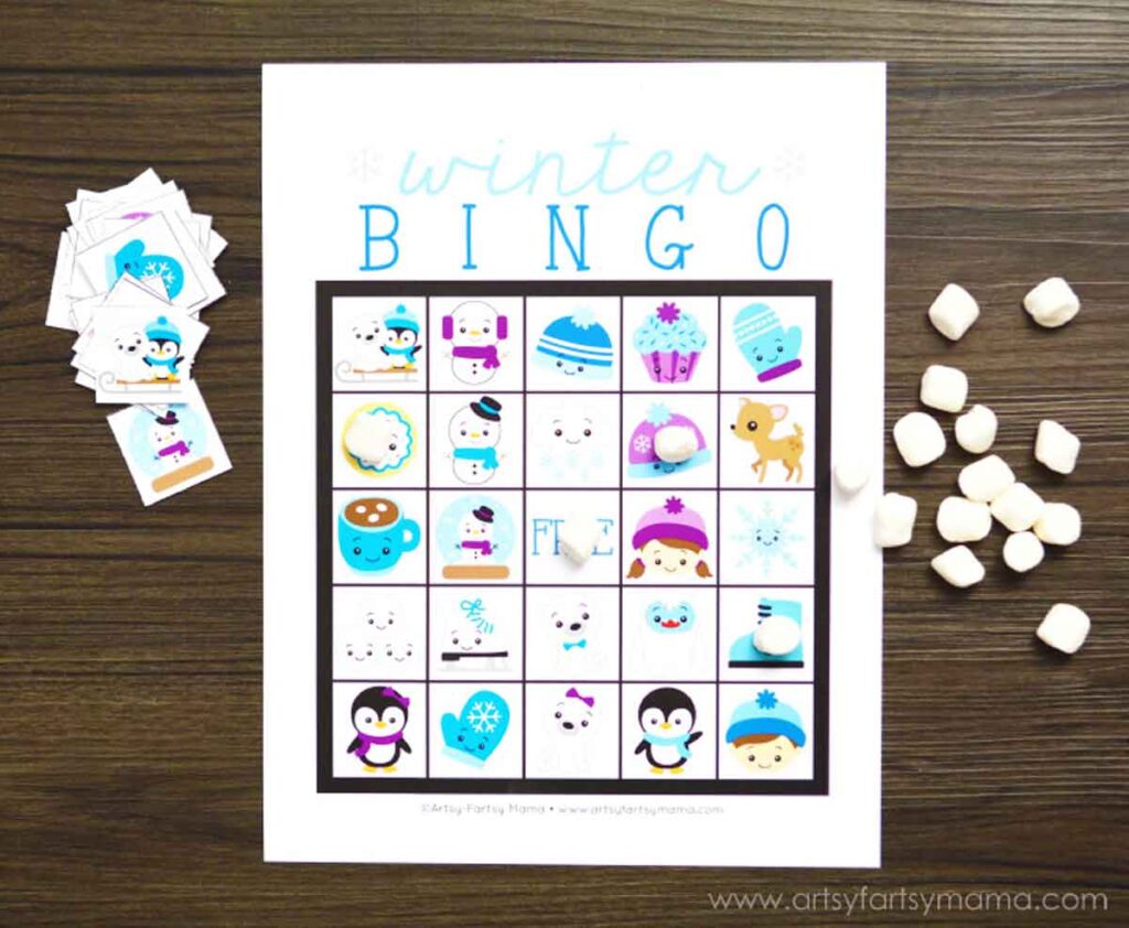 A printable winter games bingo card featuring charming illustrations of penguins, snowflakes, and hot cocoa. Small cards and marshmallows accompany it on a wooden surface, perfect for cozy game nights.