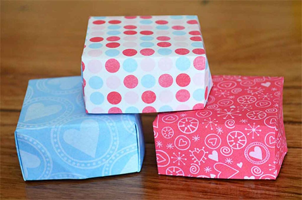 Three small gift boxes on a wooden surface, perfect as Valentine box ideas for school. One is wrapped in blue and white heart patterns, another in red and pink geometric designs, and the last features red, pink, and light blue polka dots.