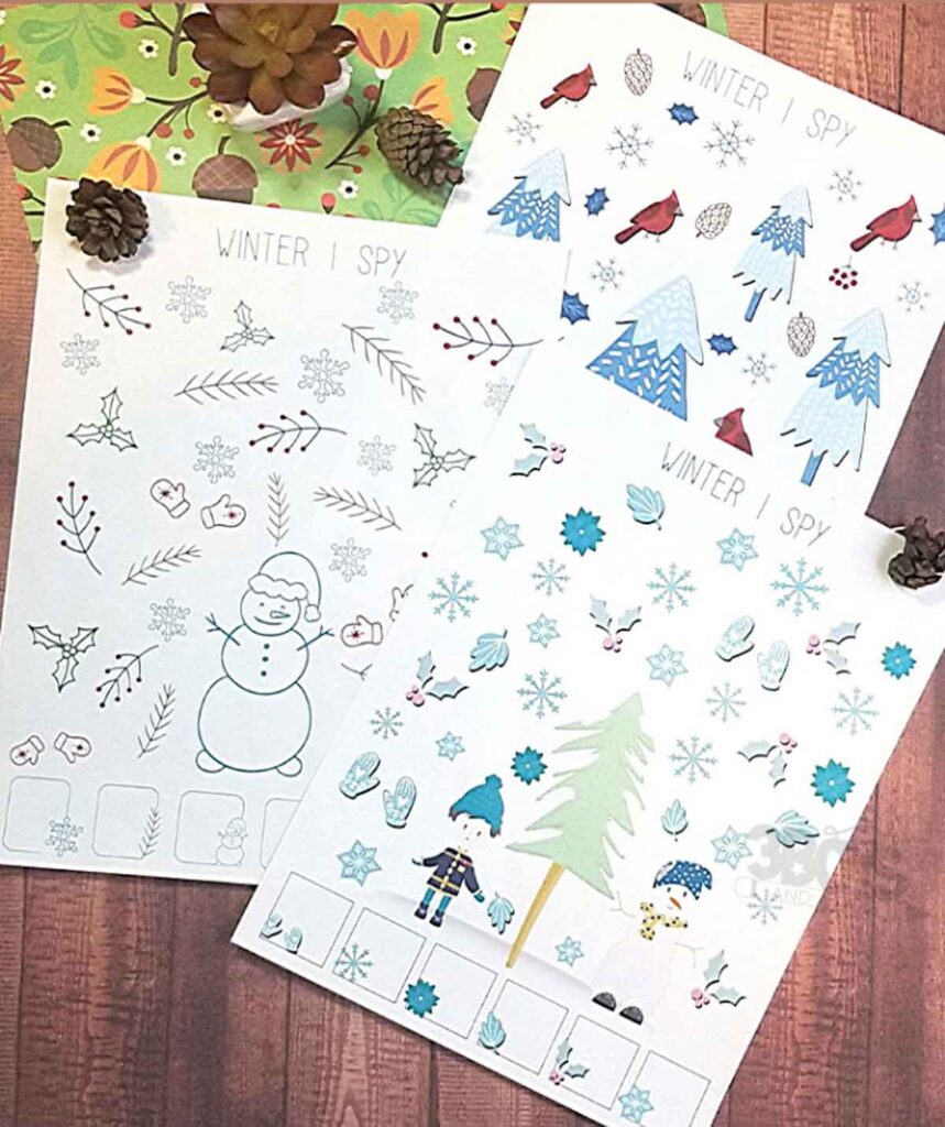 Three printable winter games rest on a wooden surface adorned with pinecones and a plant. The "I Spy" activity sheets feature snowflakes, trees, mittens, cardinals, and a snowman drawing for kids to find and count, offering festive fun.