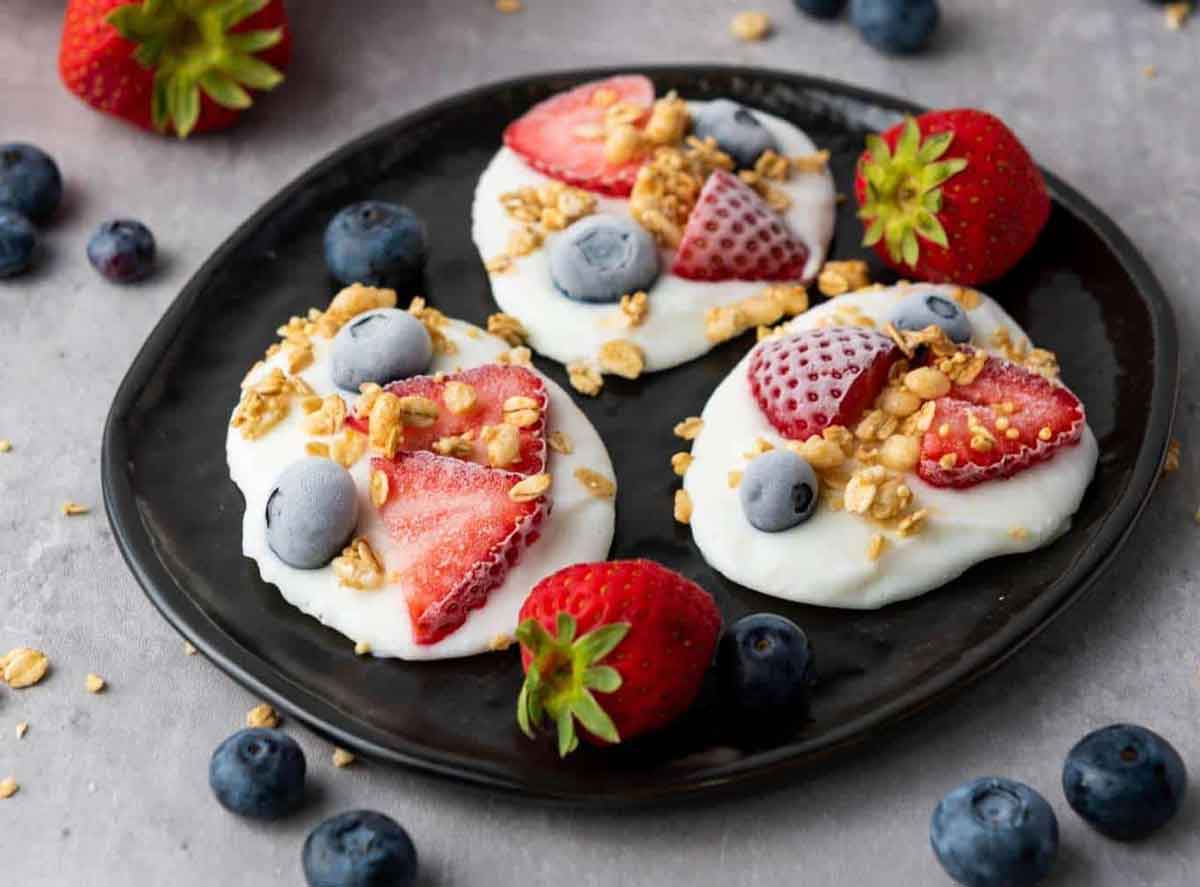 A black plate displays four yogurt clusters, garnished with sliced strawberries, blueberries, and granola, creating the perfect healthy Valentine snacks for the classroom. Fresh strawberries and blueberries are scattered around for an added touch of love.
