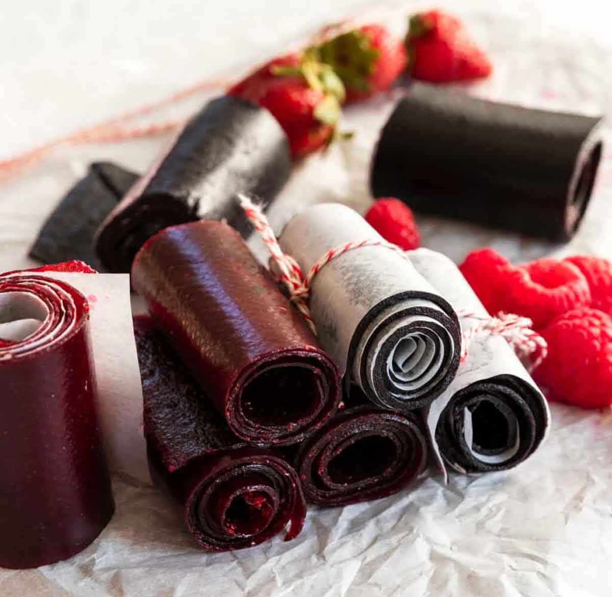 Rolled fruit leather strips in shades of red and black are displayed on a sheet of white parchment paper. Perfect as healthy Valentine snacks for the classroom, some are wrapped in white paper and tied with red and white twine, surrounded by fresh raspberries and strawberries.