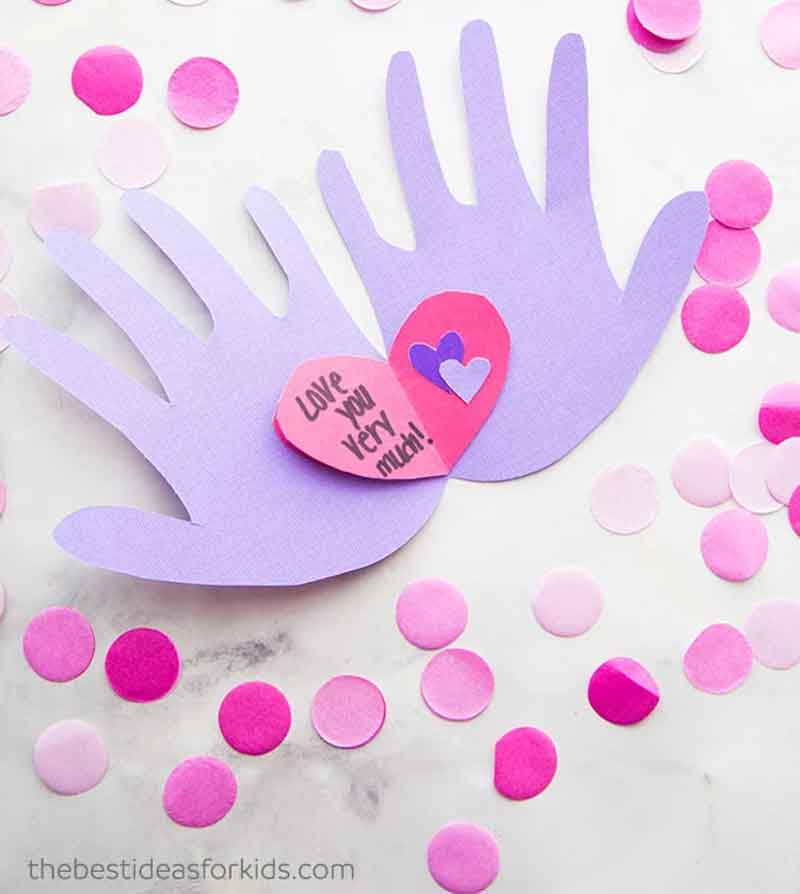 A handmade card features two purple paper hands with a pink heart in the center, bearing the message "Love You Very Much." Pink and purple confetti dots are scattered around on a white background.