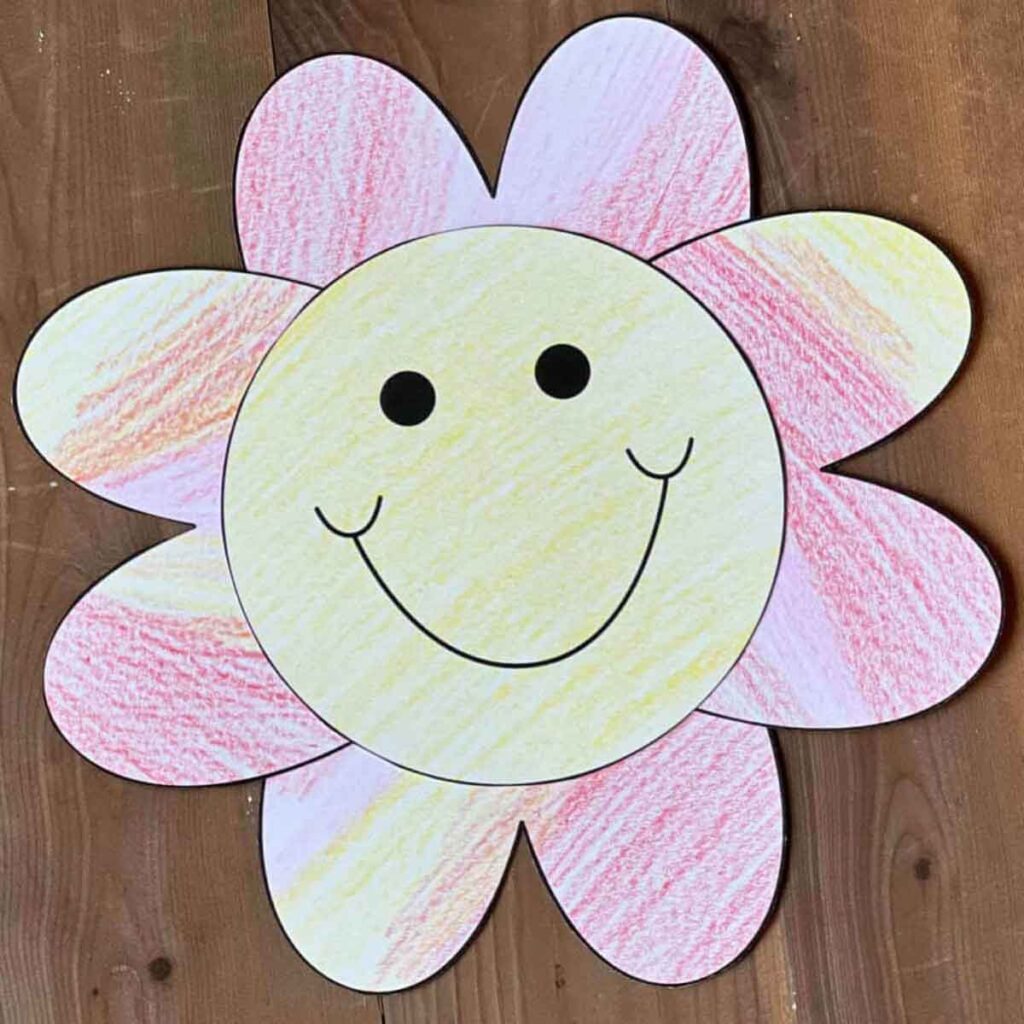 A colorful paper flower with a yellow and pink crayon-smudged pattern, perfect for Valentine paper crafts. It features a large smiling face with two black eyes on the petals, sitting on a wooden surface.