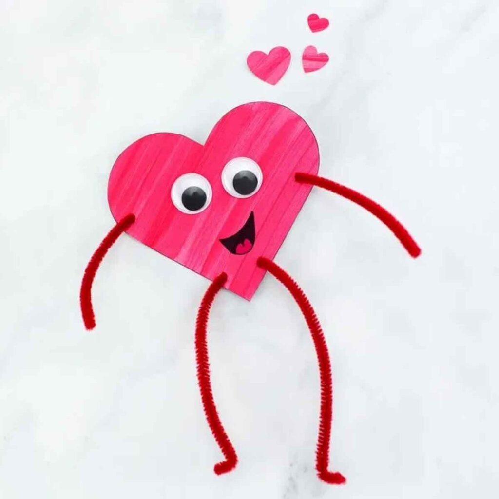 A pink heart with googly eyes and a smiling face boasts red pipe cleaner arms and legs, capturing the charm of valentine paper crafts. Three smaller pink hearts float above it on a light background.